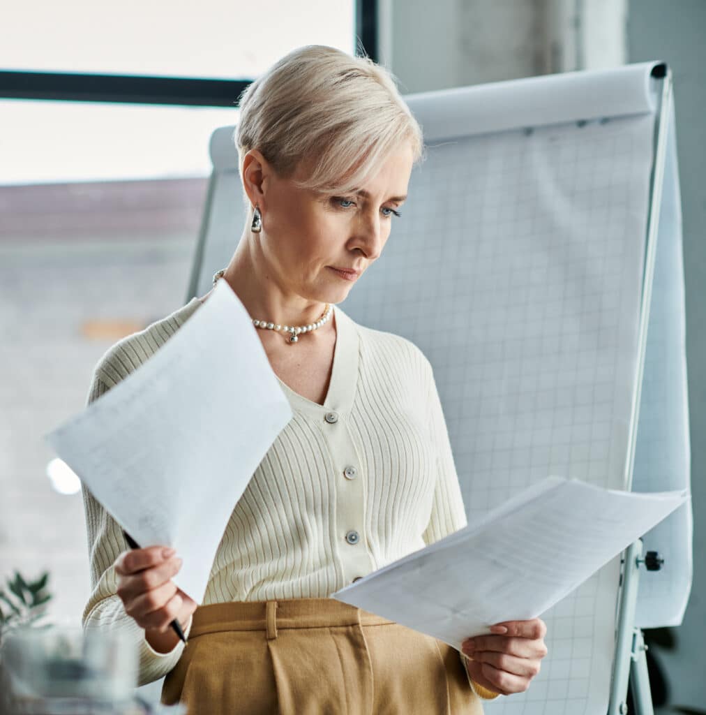 A middle-aged businesswoman with short hair holds a piece of paper in front of a computer screen.